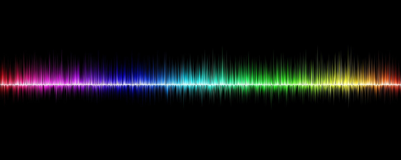#PraCegoVer Audiodescrição resumida: Imagem retangular com fundo preto. Localizado no centro, há uma representação de uma onda sonora. Nela, da esquerda para direita, estão as cores em degradê: vermelha, rosa, azul, verde, amarela e laranja.