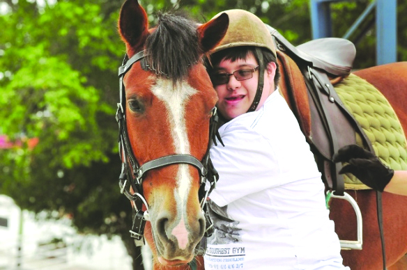 #PraCegoVer Fotografia. Um menino com síndrome de down abraça um cavalo marrom. O menino possui pele branca, usa uma camiseta branca, um óculos de grau e um chapéu equestre. Desfocado, ao fundo, há uma vegetação.
