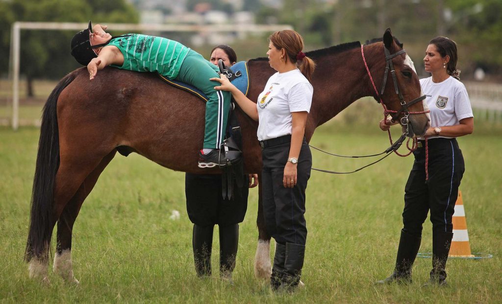 #PraCegoVer Fotografia de uma sessão de Equoterapia em um campo aberto. Um menino está deitado de braços abertos em cima de um cavalo marrom. Ele possui pele branca, usa uma camiseta e uma calça verde e um chapéu equestre. Ao redor do cavalo, há três mulheres que auxiliam o menino. Elas possuem pele branca, cabelos castanhos, usam camiseta branca, calça preta e bota de montaria preta.