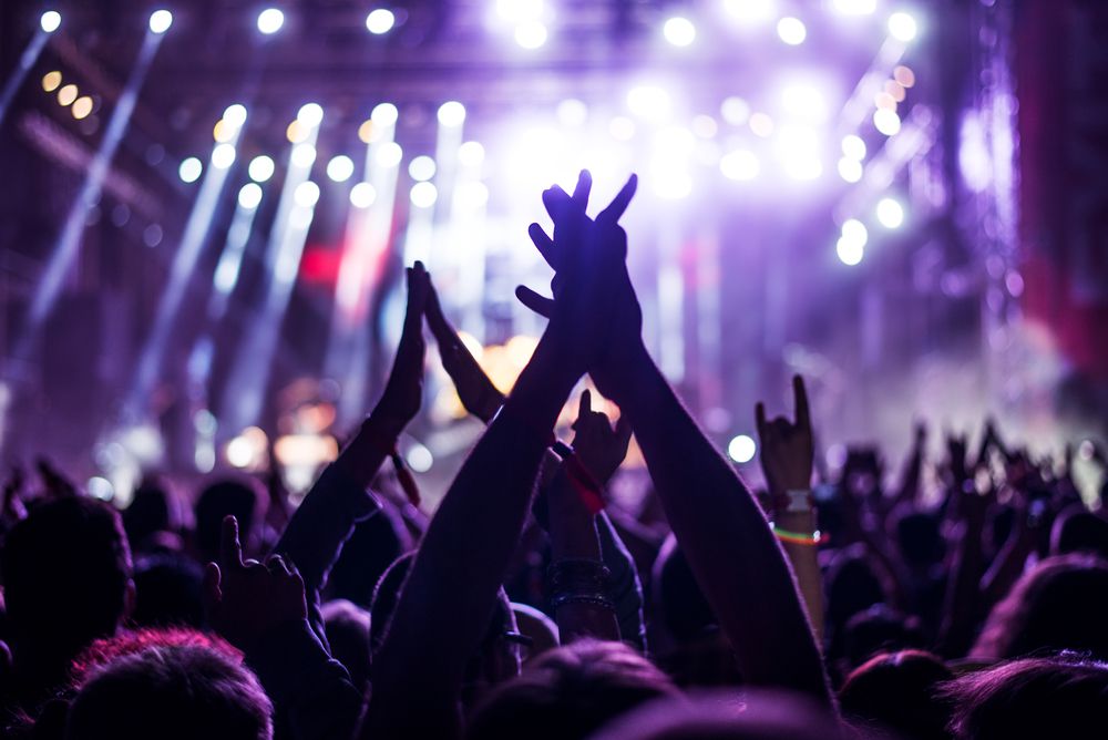 #PraCegoVer Audiodescrição Resumida: Fotografia de um show musical. Nela, há diversas pessoas com os braços para cima. Desfocado, ao fundo, há um palco com holofotes que transmitem luzes roxas.