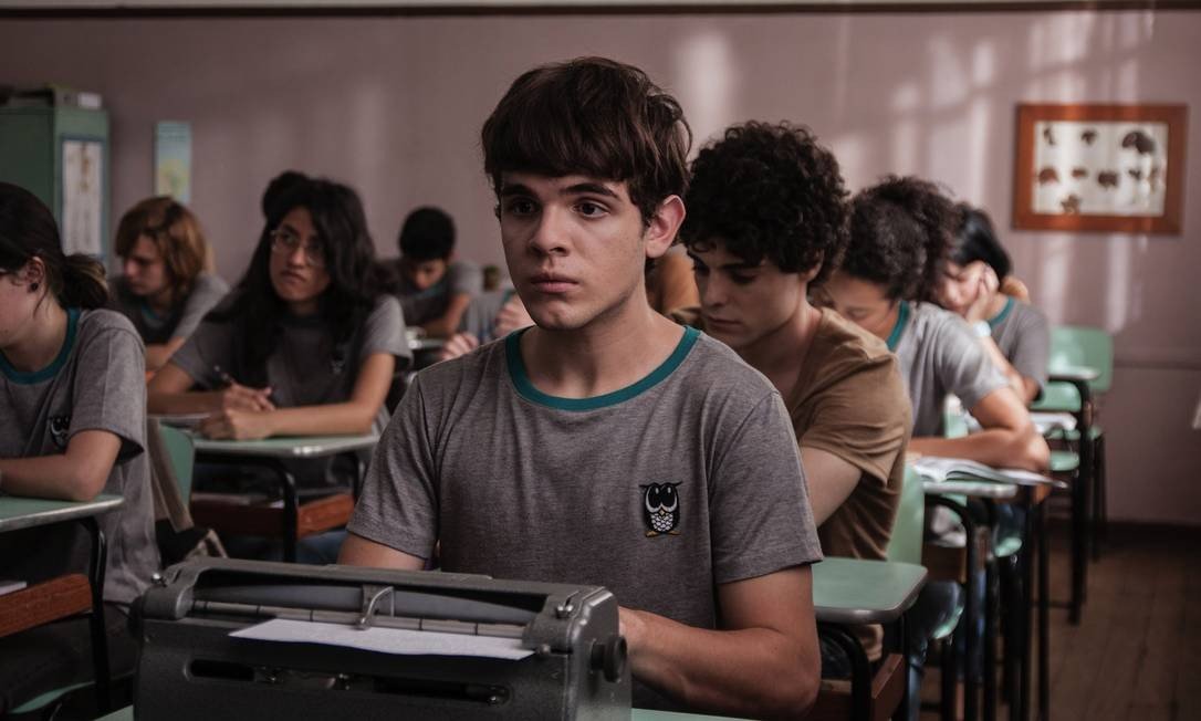 #PraCegoVer Audiodescrição resumida: Fotografia do filme “Hoje eu quero voltar sozinho”. Em uma sala de aula, há meninas e meninos sentados em cadeiras, debruçados sobre mesas enfileiradas. Sentado à frente, há um menino de pele branca, cabelos e olhos castanhos. Ele usa uma camiseta cinza com um pequeno desenho de uma coruja à direita. Em cima de sua mesa há uma máquina de escrever em braile.