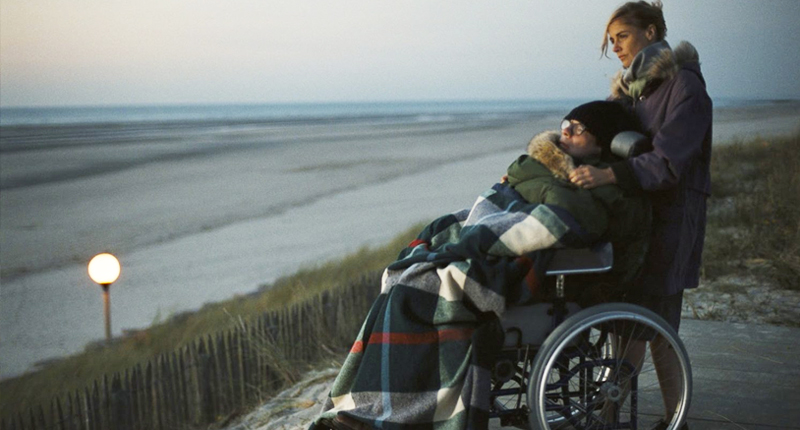 #PraCegoVer Audiodescrição resumida: Fotografia do filme “O Escafandro e a Borboleta”. Em uma praia, uma mulher está em pé atrás de um homem, que está sentado em uma cadeira de rodas. A mulher possui pele branca, cabelos loiros presos e usa roupas de frio. O homem usa um óculos de grau, uma touca e está coberto com uma manta xadrez. Ao fundo há um mar e um céu acinzentado.