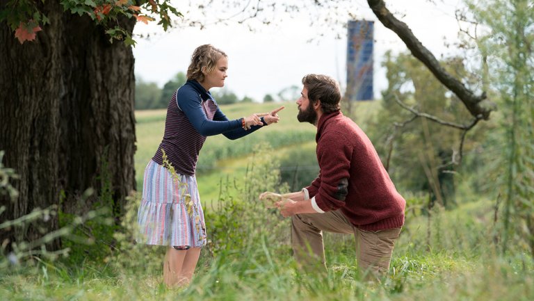 #PraCegoVer Audiodescrição Resumida. Foto do filme "Um Lugar Silencioso". Em um campo aberto, uma menina e um homem estão frente a frente. Ela faz um sinal com ambas as mãos. O homem está ajoelhado e olha fixamente para a menina.