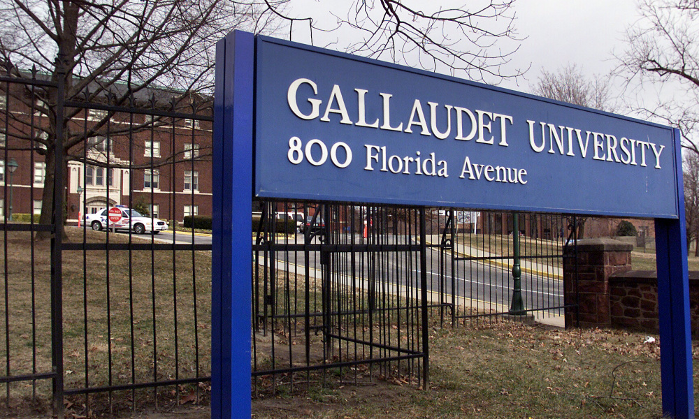 #PraCegoVer Audiodescrição resumida: Fotografia da fachada da Universidade Gallaudet. Presa ao chão, há uma placa em tom de azul com o nome da universidade. Atrás da placa há uma grade. Ao fundo da foto, há um prédio em tom de marrom e um carro estacionado na frente.