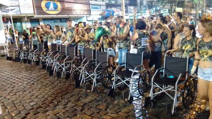 #PraCegoVer Audiodescrição resumida: Fotografia tirada em uma área externa. Em uma calçada de paralelepipedos há diversas pessoas uniformizadas e enfileiradas. À frente da primeira fileira estão postas cadeiras de rodas.