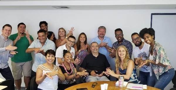 #PraCegoVer audiodescrição resumida: Em uma sala branca, quinze integrantes da equipe de tradutores de libras do samba-enredo de São Paulo estão juntas e sorriem para a foto. A maioria faz o mesmo sinal de mão, enquanto outros fazem apenas gestos.