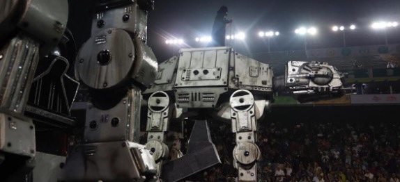 #PraCegoVer audiodescrição resumida: Fotografia de um carro alegórico na temática de Star Wars. Ao fundo, há uma plateia. Ao centro está o carro alegórico em formato de nave espacial. Acima da nave está o personagem Darth Vader. À esquerda, dois suportes metálicos grandes com um círculo no meio. No centro da imagem, há uma parte metálica em formato de cão robótico.