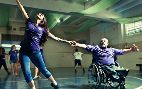 #PraCegoVer Audiodescrição resumida: Fotografia registrada em uma quadra de esportes. No centro da imagem, há uma mulher em pé e um homem cadeirante de mãos dadas. A mulher veste uma calça jeans e o homem uma bermuda verde. Ambos vestem uma camiseta roxa e esticam os braços para cima.