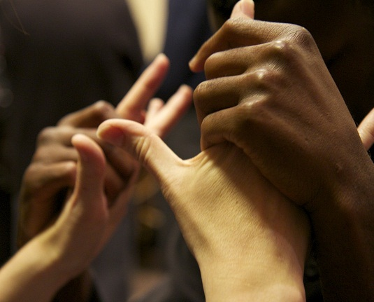 #PraCegoVer audiodescrição resumida: Fotografia. Uma mão de pele negra segura, com os dedos fechados, uma mão de pele branca, que está com a palma aberta. Ao fundo, reflexo desfocado das mãos.