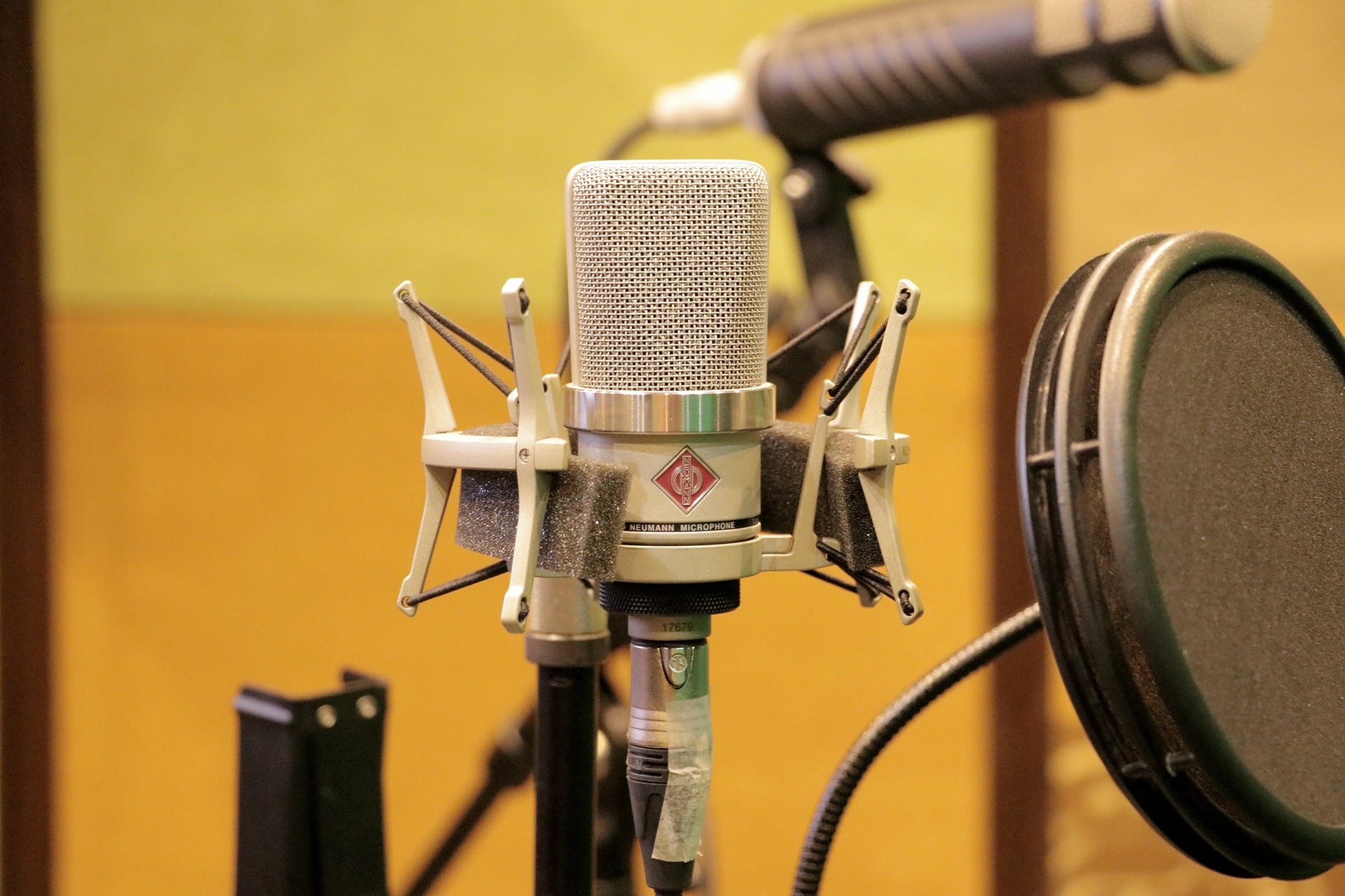 #PraCegoVer Audiodescrição resumida: Fotografia em um estúdio. Ao fundo,parede amarela e um microfone em desfoque . À Frente, a direita, um filtro circular preto de microfone. No centro, um microfone condensador prateado.